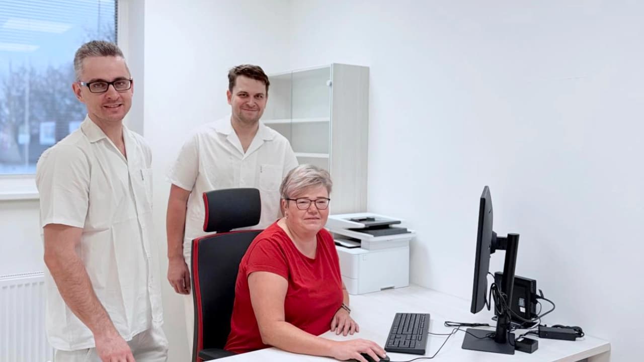 Tým lékařů a sestry v bílých a červených oděvech usmívající se v moderní světlé kanceláři u pracovního stolu s počítačem.