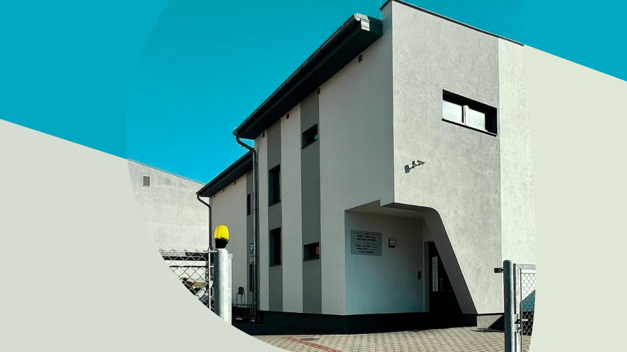 Moderní dvoupatrová bílá budova s šedými svislými pruhy, zasazená do grafického rámce s modrým nebem a krémovými plochami.